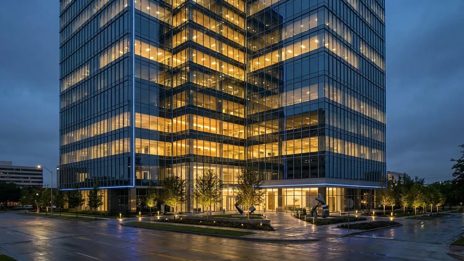 A modern Class A glass office tower at dusk in North Dallas with warm interior lighting and a blue-toned sky