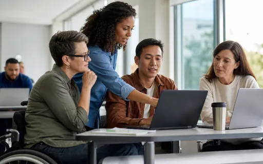 Four coworkers, including a wheelchair user, collaborate around two laptops in a bright office